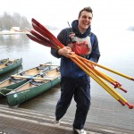 A young man smiling while carrying rowing equipment.